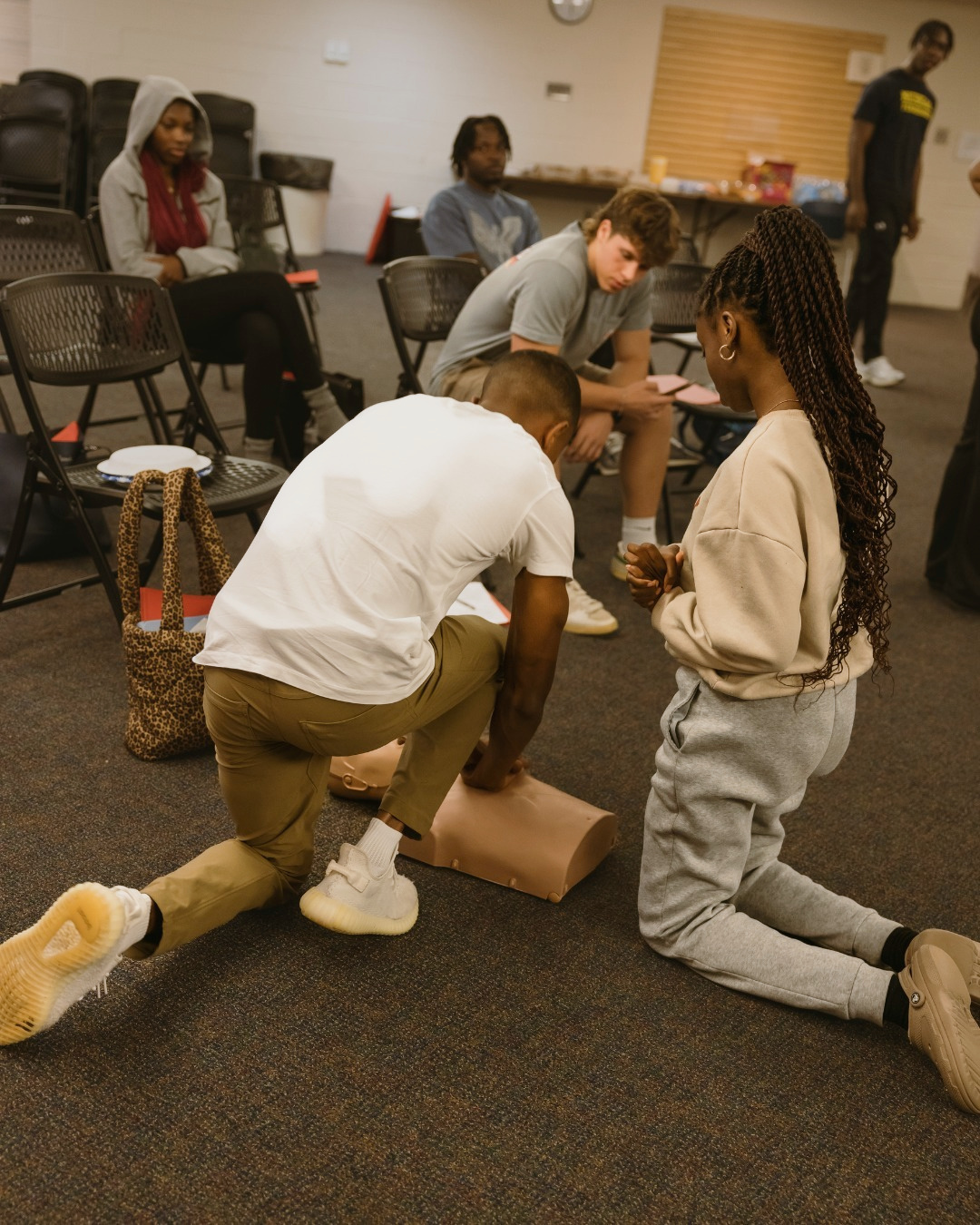 A young community member practices CPR on a training manikin while other participants watch during a GoodWorks class
