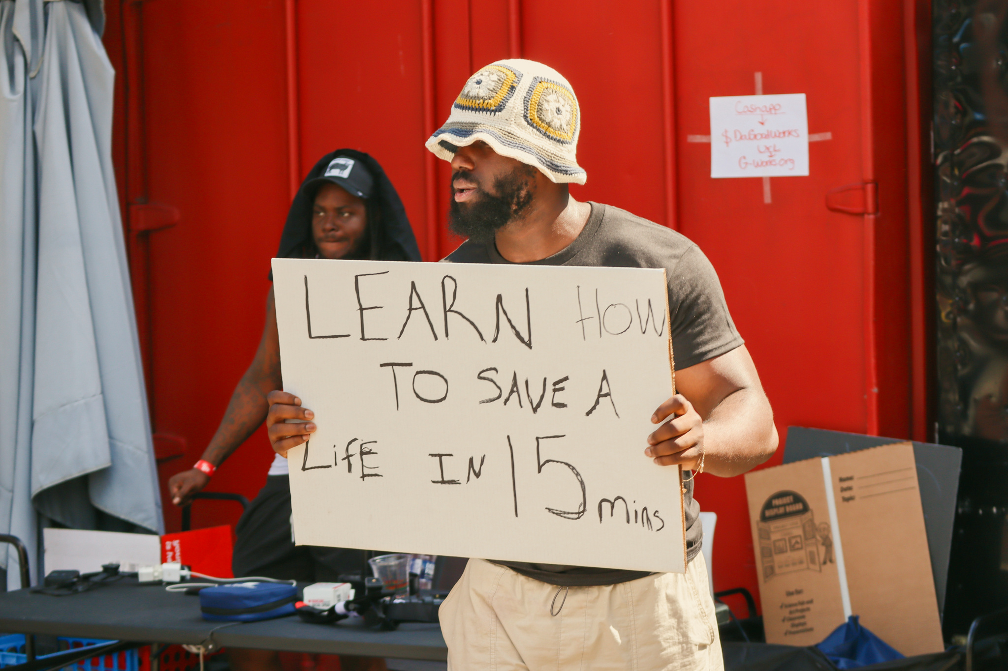 A GoodWorks volunteer holds a sign reading Learn how to save a life in 15 mins at a community event