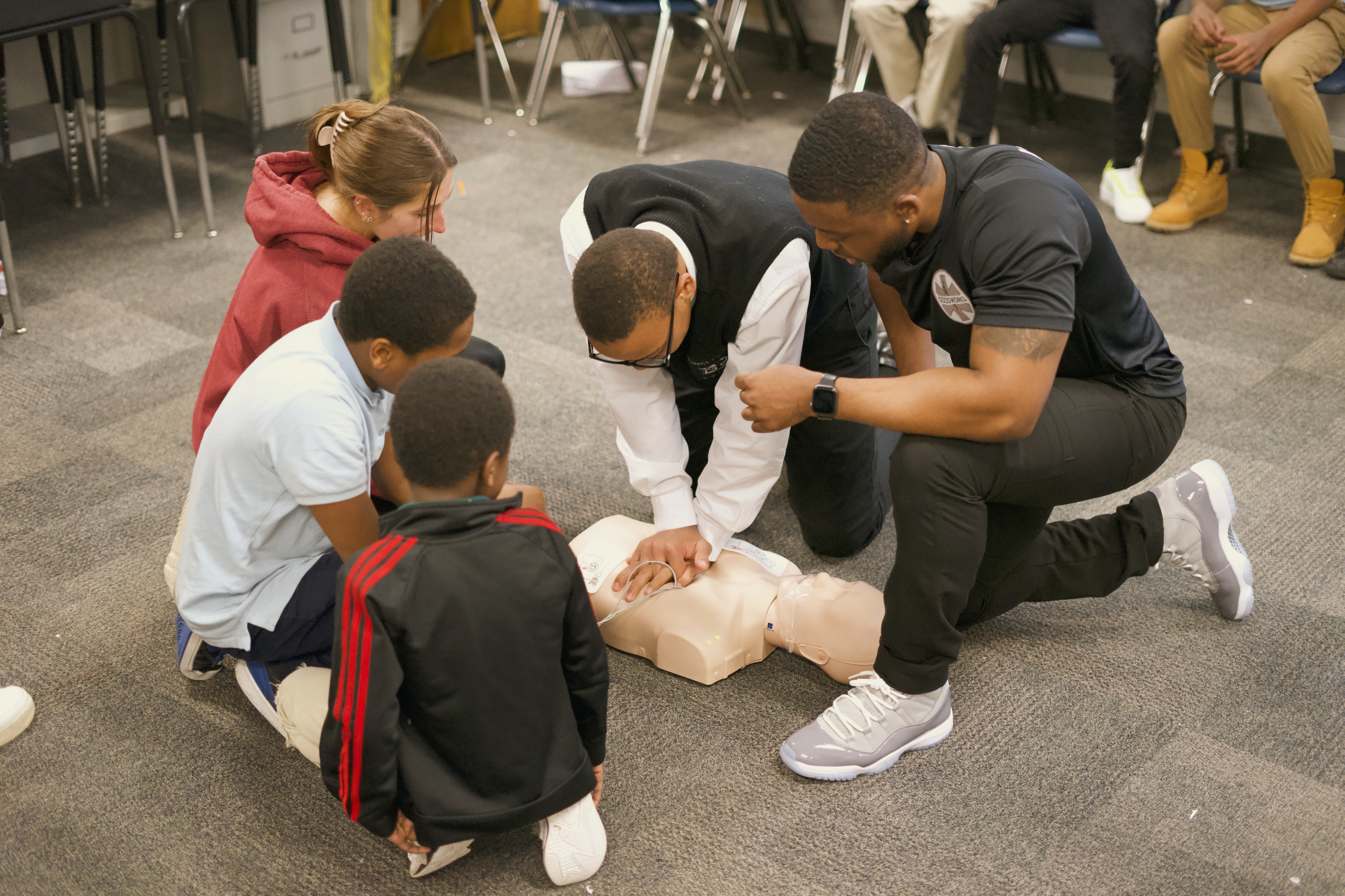 An instructor in a black shirt kneels beside school-age students practicing CPR on a floor manikin