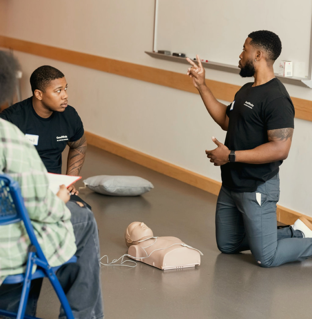 The GoodWorks founder kneels beside a CPR manikin to teach two seated community class attendees